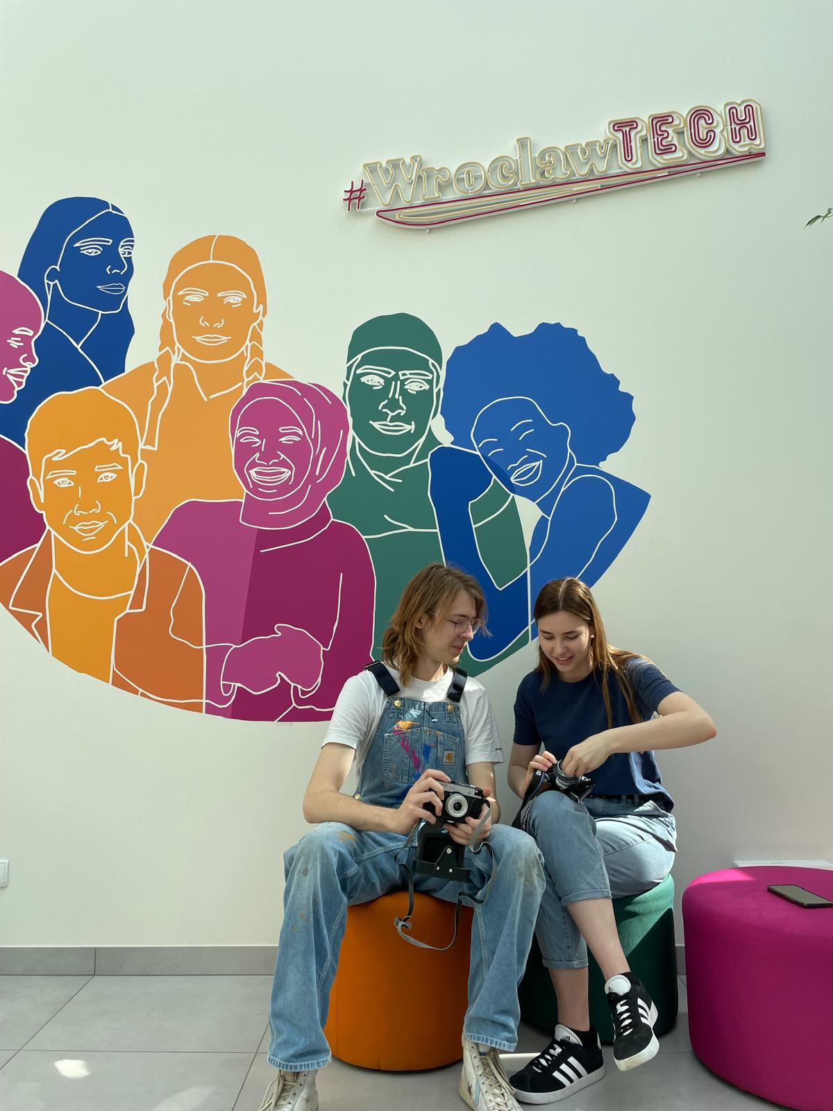 Two students standing in front of a mural depicting people of diverse backgrounds.